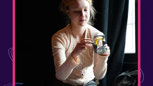 Auf dem Bild ist Sophia Eisenhut zu sehen, wie sie ein Legobauwerk in der Hand hält. Oben links steht Litnova. Unten rechts steht Prosanova.